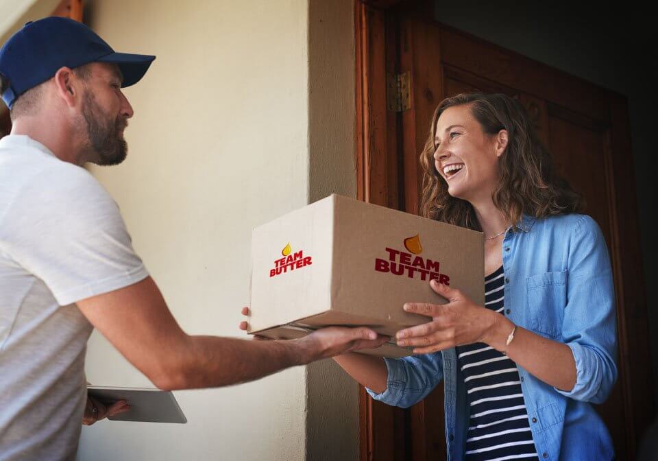 A delivery man hands a woman a box of frozen cookie dough with the Team Butter label on the side.